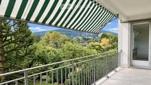 Balcony with metal railing, green and white striped awning, overlooking a lush green landscape with trees and distant hills.