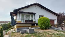 A two-story house with a balcony, surrounded by a garden area featuring a grey outdoor sofa, a small table, and an umbrella. The garden includes a large, well-maintained bush and a wooden fence.