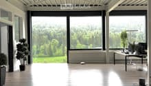 Open-plan office space with wooden flooring, large windows overlooking a green landscape, a desk with a laptop, and potted plants.
