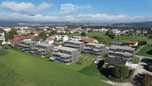 Aerial view of a residential area with modern apartment buildings, green lawns, and surrounding town with houses and fields.