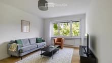 Modern living room with a grey sofa, orange armchair, black coffee table, TV stand, and a large window with white curtains.