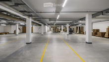 Large, empty warehouse or industrial space with concrete floors, white walls, and visible storage racks and pallets.