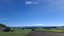 A panoramic view of a lush green field with scattered trees and distant mountains under a clear blue sky.