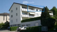 A modern, multi-story residential building with white exterior walls, multiple balconies, and a well-maintained garden area. The building features large windows and a clean, contemporary design.