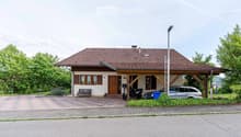 Single-story house with a tiled roof, wooden entrance, and covered porch area. The house has multiple windows with shutters, a bench near the entrance, and a driveway with parked vehicles including a car and motorcycles.