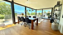 Bright dining area with wooden floor, large windows offering a scenic mountain view, dining table with chairs, decorative shelf, and potted plants.