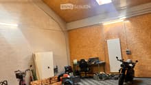 A spacious workshop or garage with a slanted wooden ceiling, exposed brick wall, and checkered floor. Contains a desk with a chair, a motorcycle, a bicycle, a fan, a radiator, and various storage items.