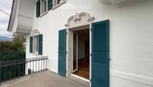 Balcony with metal railing, green shutters, and a view of the house entrance and surrounding greenery.