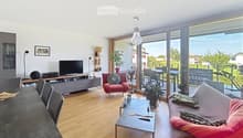 Modern living room with wooden flooring, large windows offering a view, a sofa, armchair, coffee table, dining table, TV, bookshelf, and potted plants.