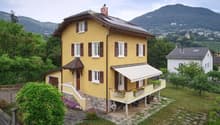 Two-story yellow house with white-framed windows, brown shutters, and a balcony with flower boxes. Solar panels on the roof and a scenic mountain view in the background.