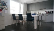 Modern kitchen with grey cabinets, white dining table, four black chairs, and various appliances including a microwave and a faucet.