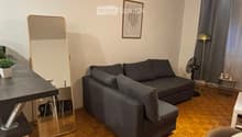 A modern living room with wooden flooring, a grey sofa, a small table, a mirror, a fan, and a lamp.