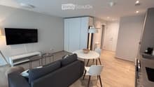 Modern living room with a grey sofa, coffee table, dining table, chairs, and a television mounted on the wall. The room features light wooden flooring and white cabinets.