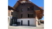 Modern two-story building with wooden facade, large windows, and balconies. The ground floor has a glass entrance and a small seating area.