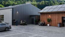 Modern building with grey and wooden facades, solar panels on the roof, outdoor seating area with picnic tables, and a barbecue area.