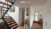 Modern hallway with wooden flooring, a staircase leading upwards, and artwork on the walls. A small table with a floral arrangement is placed near the staircase, and a living area with a sofa is visible at the end of the hallway.