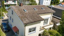 A two-story house with a tiled roof, white exterior, multiple windows, and a small garden area with potted plants.