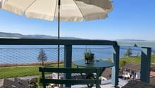Balcony with a view of a lake and surrounding landscape, featuring a round table, chairs, and an umbrella.