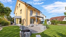 Two-story yellow house with a modern design, featuring a balcony, large windows, and a well-maintained garden with a stone fire pit.
