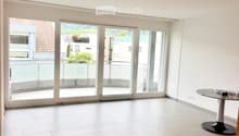 Empty room with white tiled flooring, large sliding glass doors leading to a balcony, and a small round table with books in the corner.