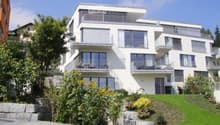 Modern white multi-story apartment building with balconies and large windows, surrounded by a well-maintained garden with sunflowers and shrubs.