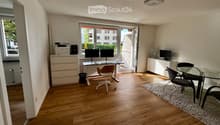 Bright, modern room with wooden flooring, large windows, and a balcony. Contains a desk with two monitors, a chair, a small table with chairs, a printer, and a fan.