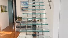Modern hallway with a sleek glass staircase featuring metal railings, wooden flooring, and contemporary decor including a small shelf unit and artwork.
