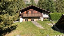 Two-story wooden house with a balcony, surrounded by trees and mountains.