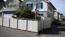 Two-story residential house with white walls, blue shutters, and a garage. A white fence surrounds the property, and a car is parked nearby.