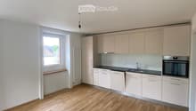 Empty kitchen with light wooden flooring, white cabinets, and a window letting in natural light. No appliances or furniture are present.