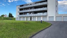 Modern multi-story apartment building with balconies and multiple garage doors, surrounded by a grassy area and a paved driveway.