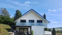 Modern two-story house with white exterior, black roof, and large windows. Features a raised ground floor, outdoor seating area, and landscaped garden.