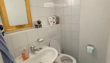 A small bathroom with a white sink, toilet, and mirror. The sink has a silver faucet and a soap dispenser. Toilet paper and a towel are visible, along with a decorative mirror and a small figurine on the windowsill.