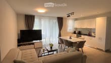 A modern, open-concept living and dining area with a light-colored sofa, coffee table, flat-screen TV, dining table with chairs, and a small kitchen area with white cabinets and a microwave.