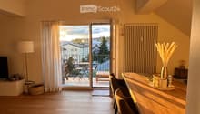 Bright, airy room with wooden flooring, large window leading to a balcony, dining table with chairs, decorative vase, and a TV on a stand.