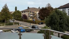 Balcony with glass railing overlooking a street with parked cars and buildings in the background.