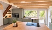 Modern kitchen with grey cabinets, wooden island, induction cooktop, double oven, and large windows overlooking a garden.