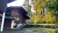 A two-story wooden house with a stone wall and steps leading to the entrance, surrounded by autumn foliage and trees.