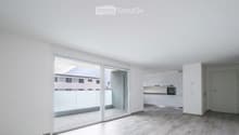 Bright, empty room with large sliding glass doors leading to a balcony, white walls, light wooden flooring, and a small adjacent kitchen area with white cabinets and appliances.