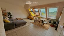Bright living room with large windows offering a scenic mountain view, featuring a grey sectional sofa, coffee table, dining area with a table and chairs, and several potted plants.