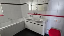 Modern bathroom with black tiled floor, white tiled walls, double sink vanity, bathtub, toilet with red seat cover, and a large mirror with decorative design.