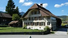 Two-story house with a brown tiled roof, multiple windows with shutters, and a satellite dish on the roof. The house is surrounded by a low stone wall and a grassy area with trees and mountains in the background.