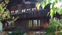 Traditional wooden house with a steep roof, multiple windows, and flower boxes on the balconies. The house is surrounded by greenery and a stack of firewood is visible in front.