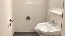 Modern bathroom with a white sink, chrome faucet, toilet, and a mirror above the sink. The walls are tiled in light beige, and the floor is covered with dark gray tiles.