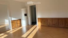 Empty room with wooden herringbone flooring, white walls, and a white cylindrical object in the center. Two doorways lead to adjacent spaces.