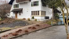 Two-story white house with green shutters, wooden balcony, and small garden area.