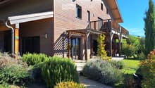 Two-story wooden house with a balcony, covered patio, and well-maintained garden with shrubs and a lawn.