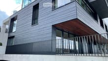 Modern multi-story building with grey brick facade, large windows, and a wooden balcony with metal railing.