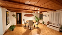 A modern dining area with a round wooden table, four chairs, potted plants, a shelf with decorative items, and a surfboard on the wall.