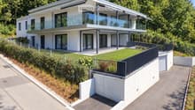 Modern two-story house with white exterior, large glass windows, and a raised ground floor. Features include a balcony, garage, and landscaped garden area.
